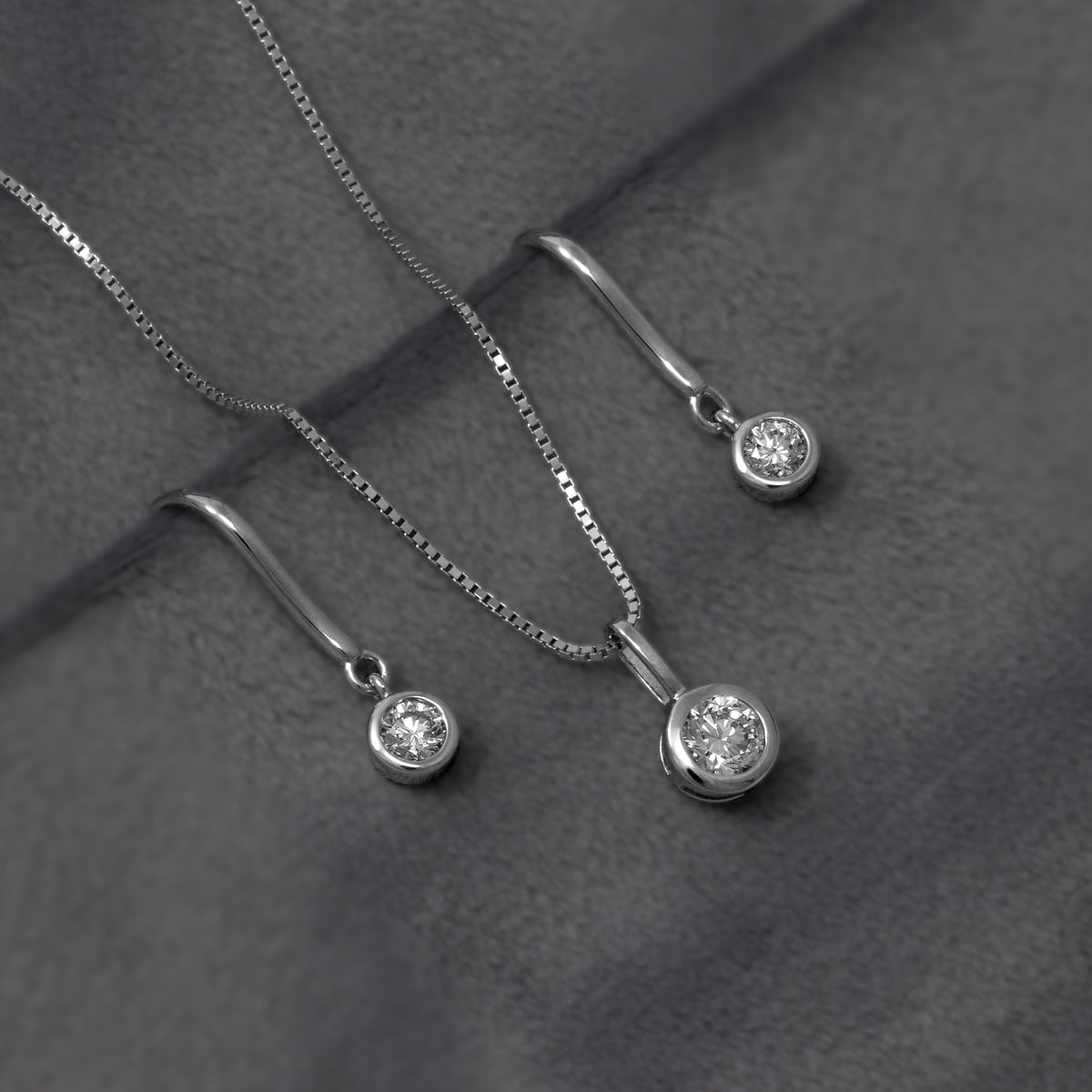 A half carat diamond pendant, and a pair of silver hook earrings each featuring a quarter carat round bezel-set lab diamond, shown against a grey background.