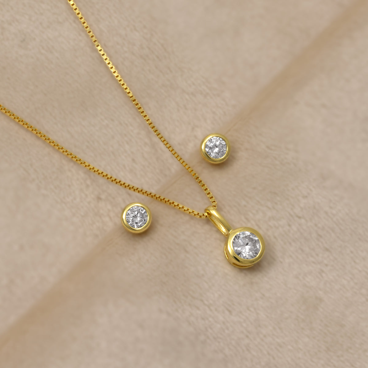 Gold vermeil necklace and matching stud earrings set with solitaire lab grown diamonds, displayed on a soft beige fabric background.