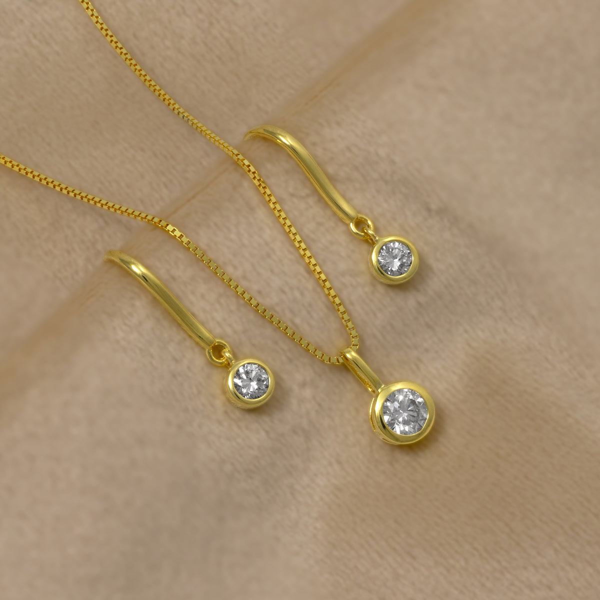 Gold vermeil pendant and drop earrings set with round bezel-set lab grown diamonds, displayed on a soft beige textured background.