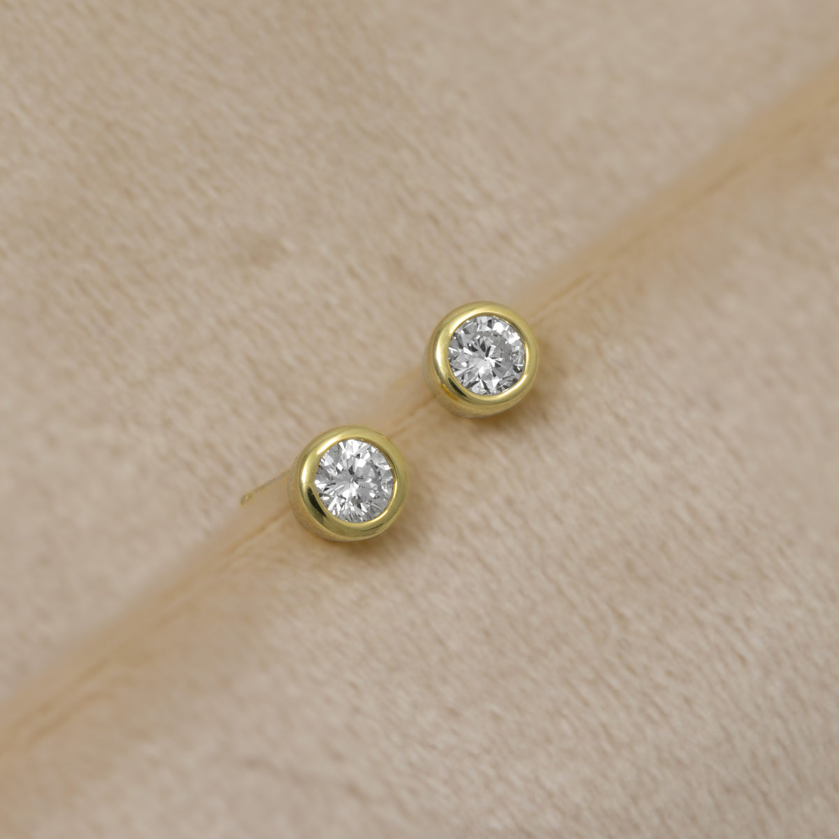 A pair of round gold vermeil stud earrings with brilliant lab grown diamonds, displayed on a textured beige surface.