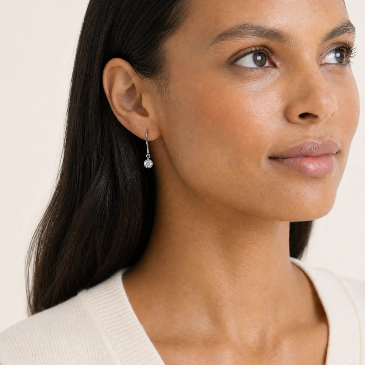 A model wears a pair of silver hook earrings, each featuring a quarter carat round bezel-set lab diamond, shown against a plain white background.