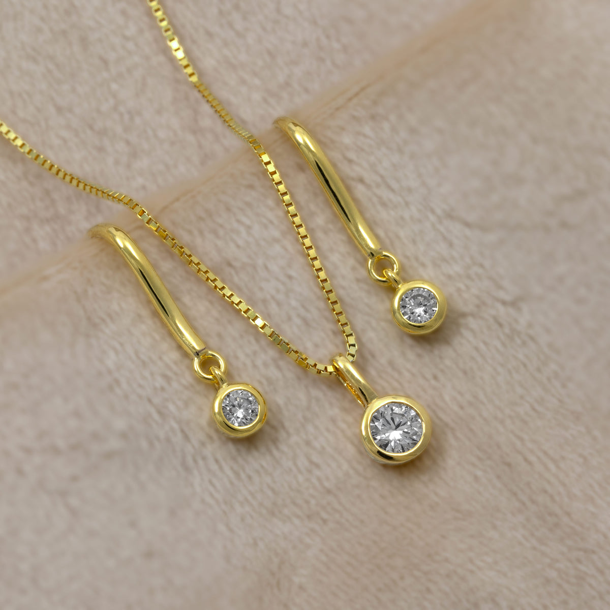 Gold vermeil lab diamond solitaire pendant and drop earrings, displayed on a soft beige textured background.