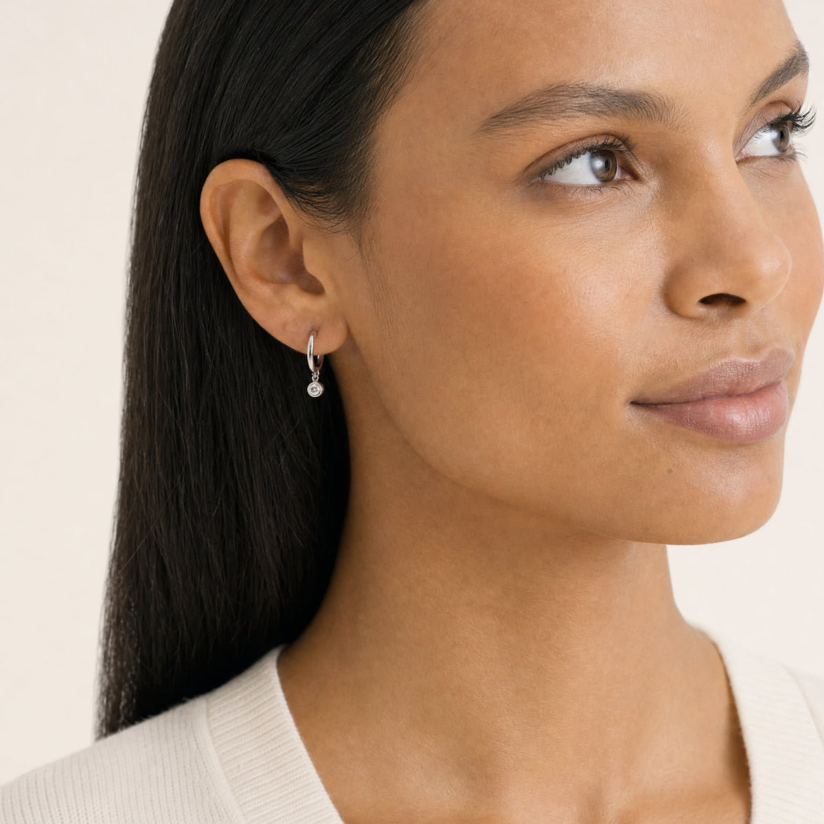 A close-up of a woman wearing a delicate silver hoop earring with a dangling lab grown diamond charm, shown against a clean light beige background.