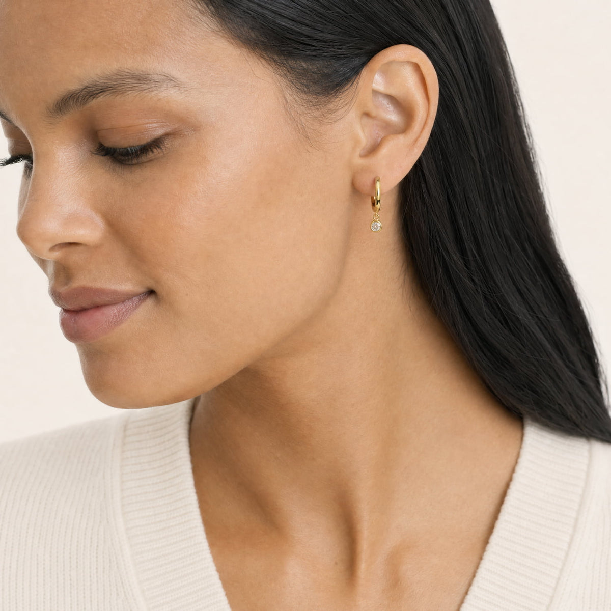 A close-up of a woman wearing a delicate gold huggie hoop earring with a lab grown diamond charm, shown against a soft beige background.