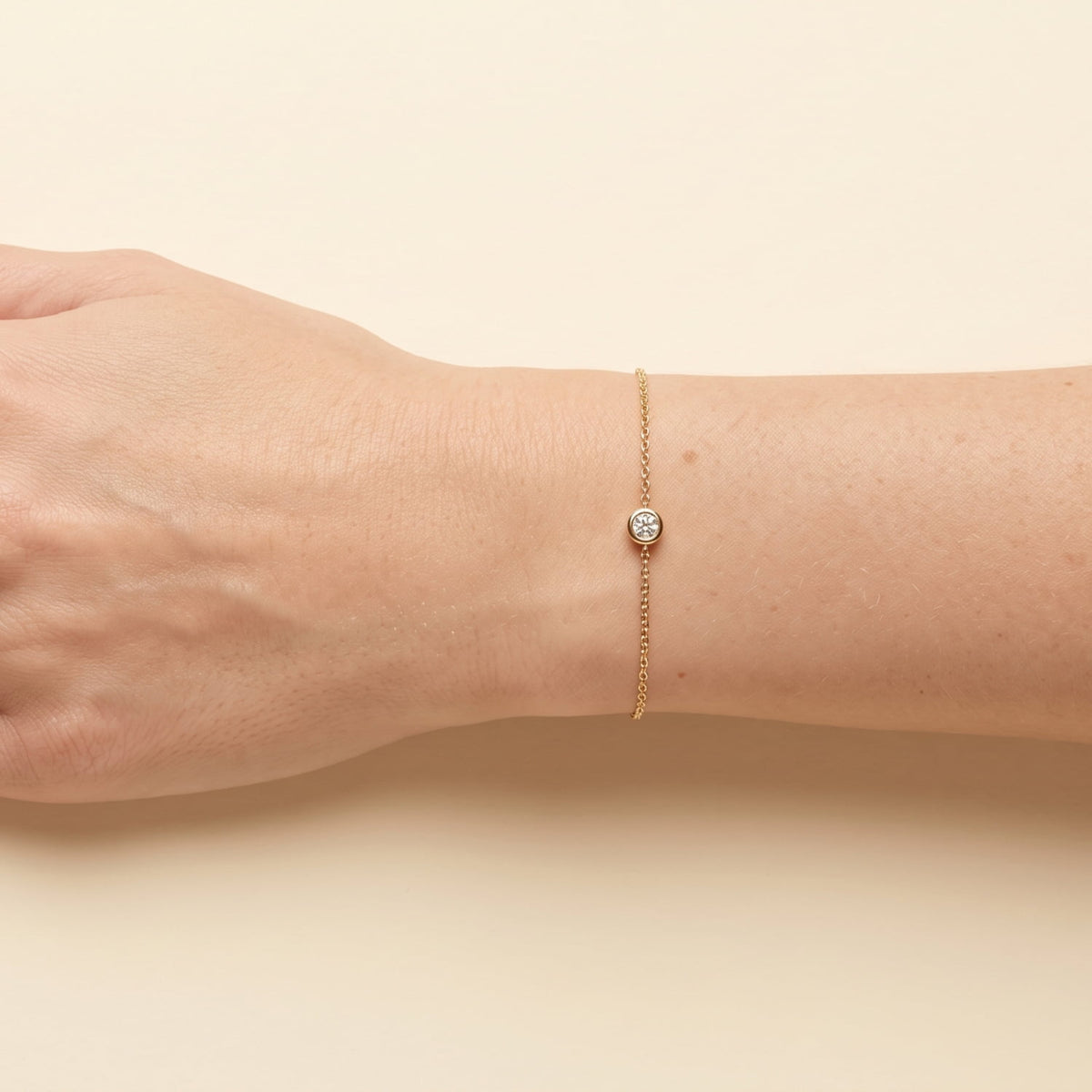 A delicate gold vermeil chain bracelet with a bezel-set lab diamond, shown worn on a person's wrist against a warm beige background.