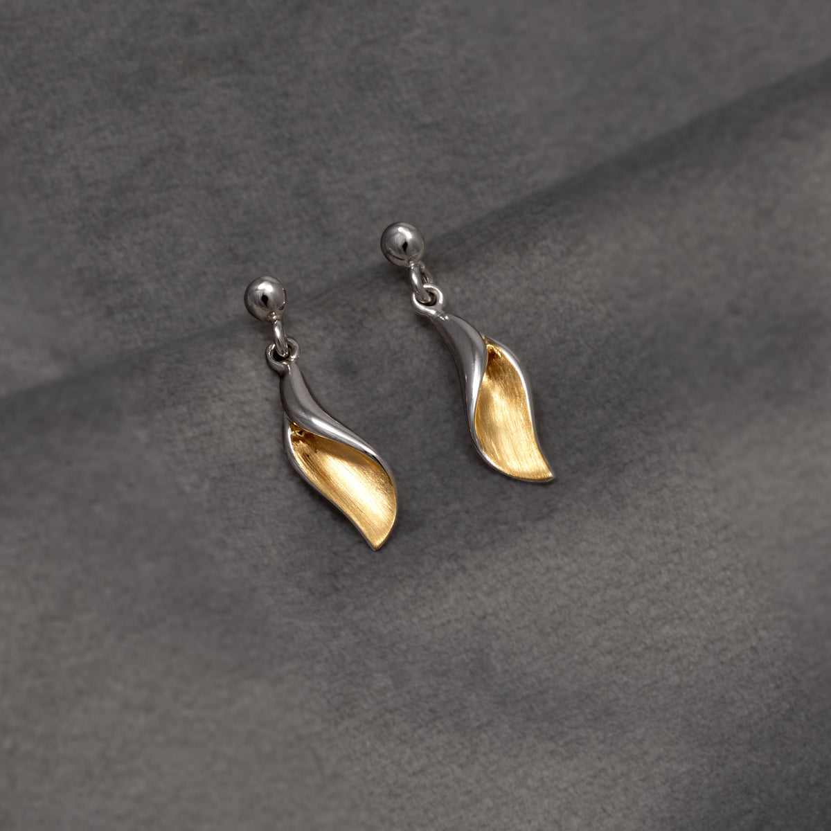A pair of silver and gold vermeil leaf-shaped drop earrings displayed on a textured dark grey fabric background.