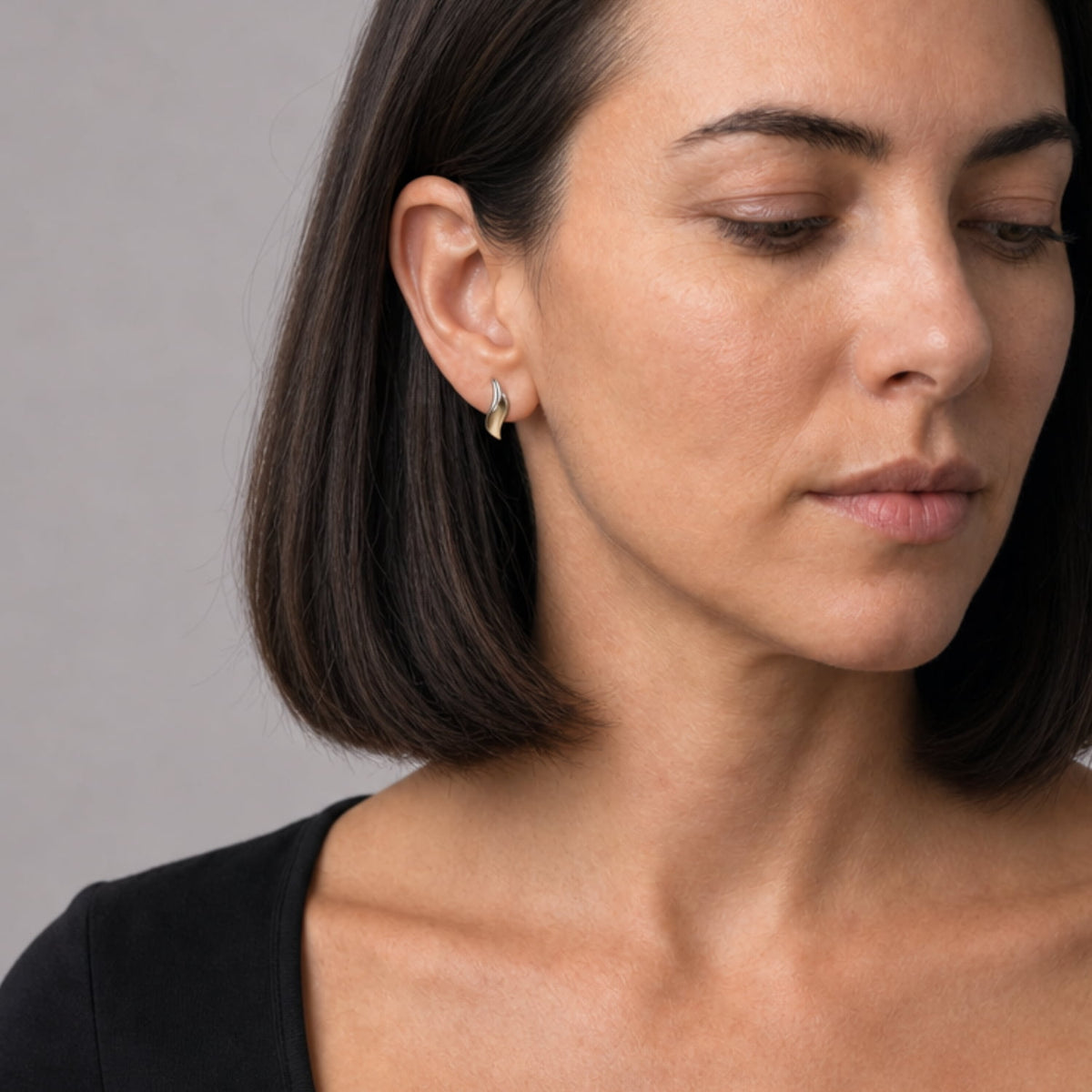 A close-up of a woman wearing a small gold vermeil and silver wave-shaped stud earring, set against a plain gray background.