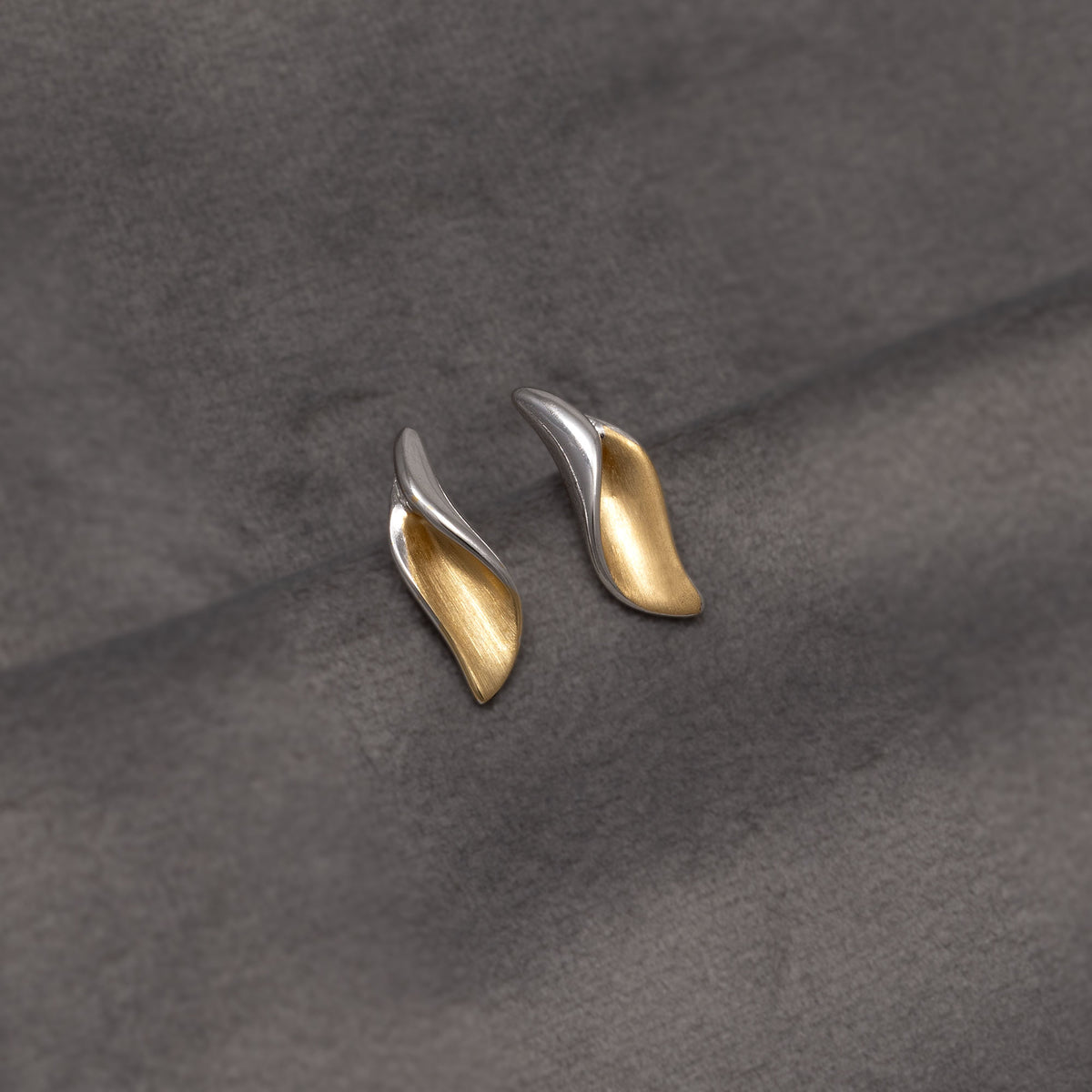 A pair of small leaf-shaped stud earrings with a two-tone finish, featuring one gold vermeil inner and one polished silver outer side, displayed on a textured dark grey fabric background.