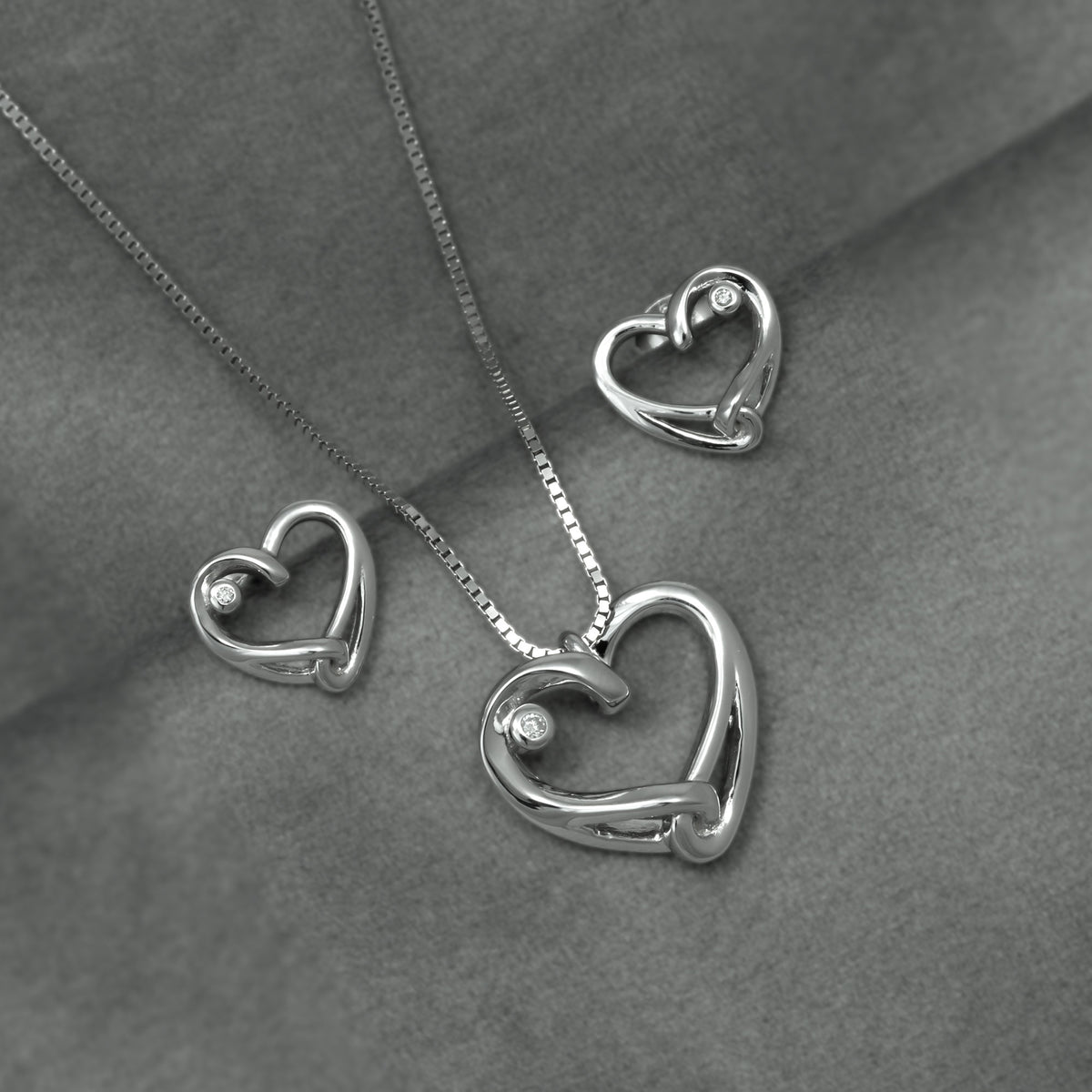 Silver heart-shaped pendant necklace and matching stud earrings with lab diamond accents on a textured gray fabric background.