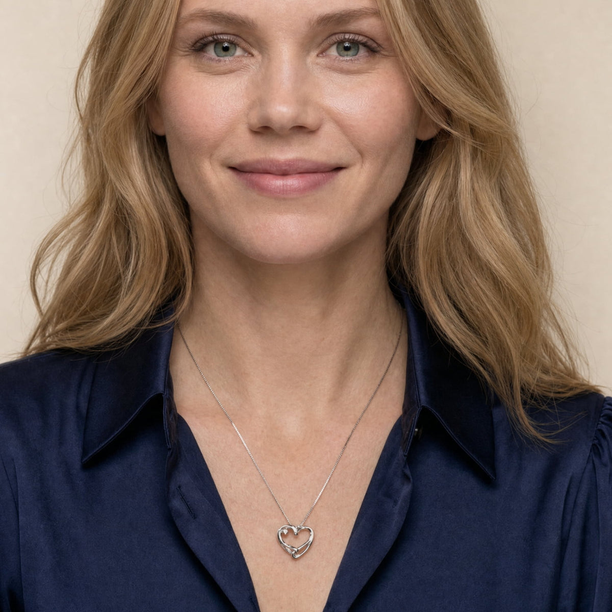 A woman with blonde hair wearing a silver heart-shaped pendant necklace and a navy blue satin shirt, photographed against a soft beige background.