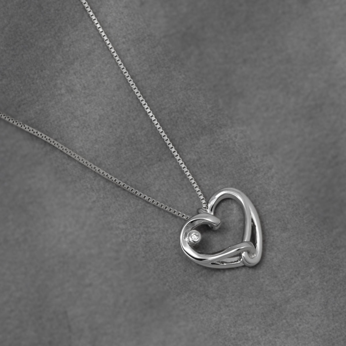 A silver heart-shaped pendant necklace with alab grown diamond accent, displayed on a textured gray background.