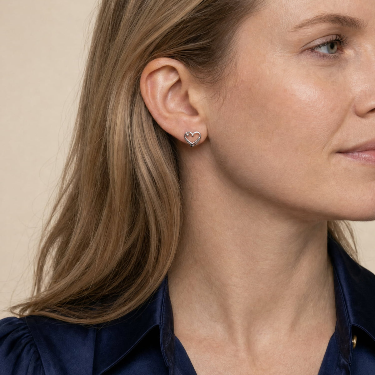 A close-up side profile of a woman with blonde hair wearing a silver heart-shaped stud earring and a navy blue satin shirt against a soft beige background.