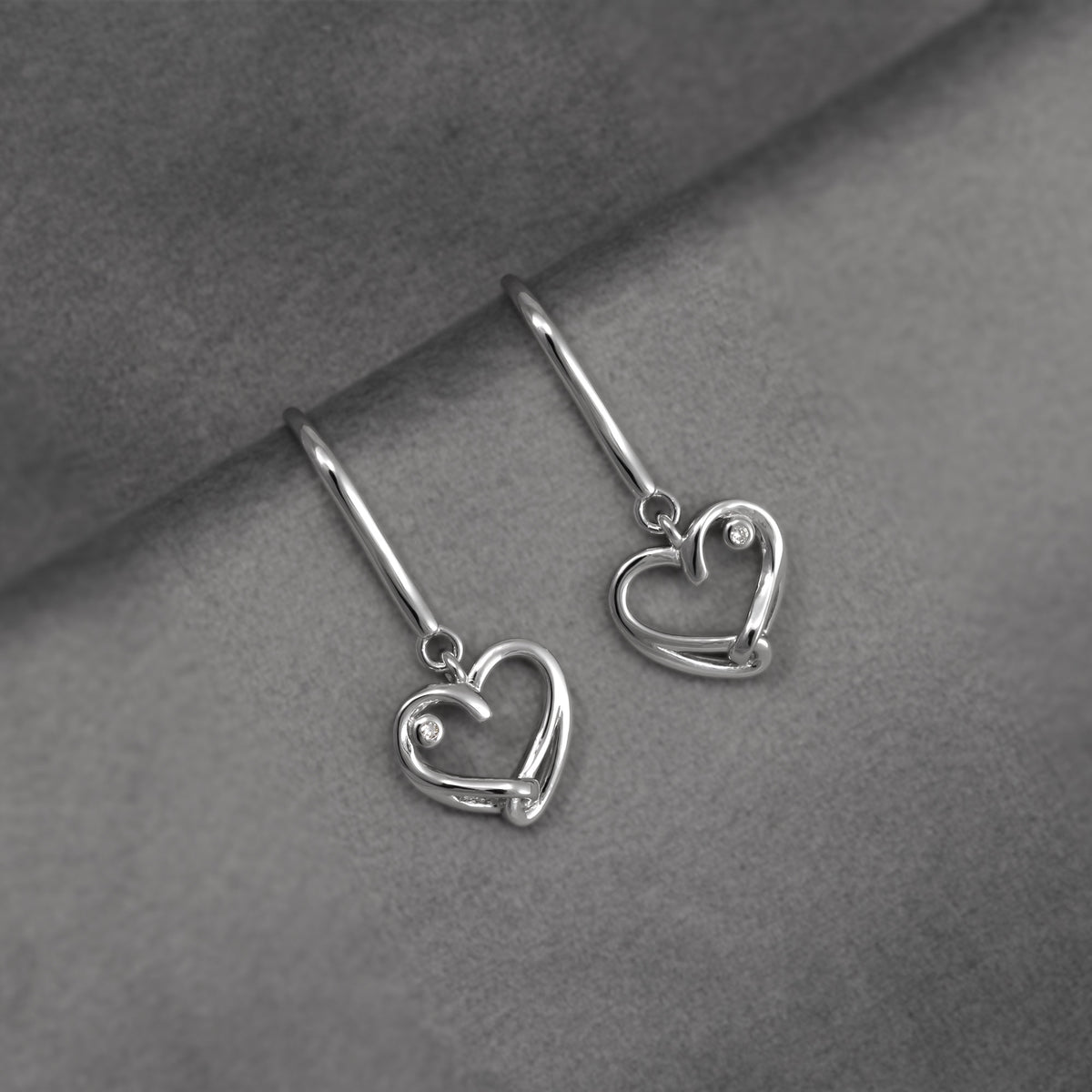 A pair of silver heart-shaped drop earrings with lab diamond accents, displayed on a textured gray surface.