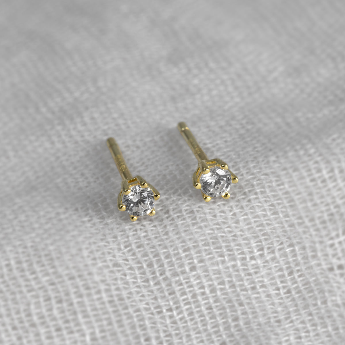 A pair of small gold vermeil stud earrings with clear round-cut cubic zirconia in a prong setting, placed on a textured white fabric background.