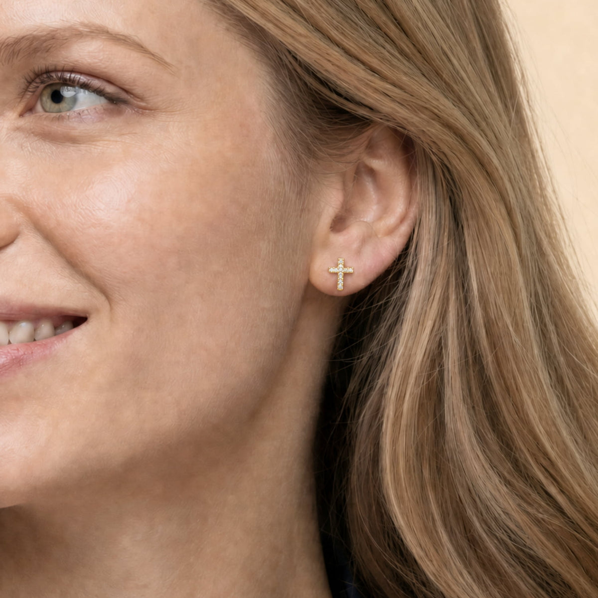 A close-up of a woman's ear wearing a small gold vermeil cross stud earring against a soft beige background.