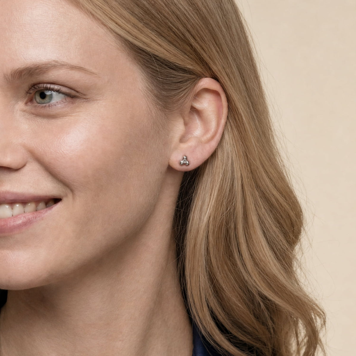A close-up of a woman's ear wearing a small silver triangular stud earring against a soft beige background.