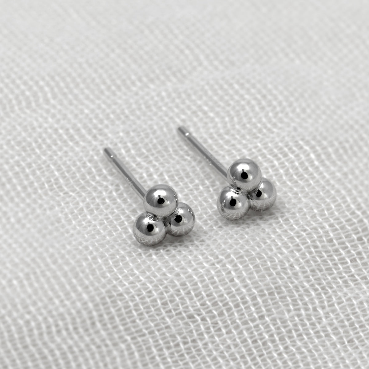A pair of silver stud earrings with three small silver beads clustered at the end of straight posts, displayed on a textured white fabric background.