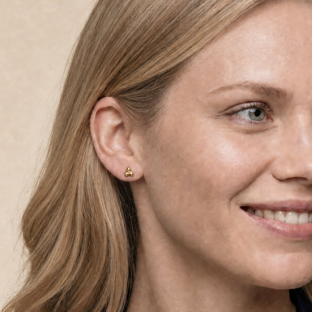 A close-up of a woman's ear wearing a small gold vermeil stud earring, set against a soft beige background.