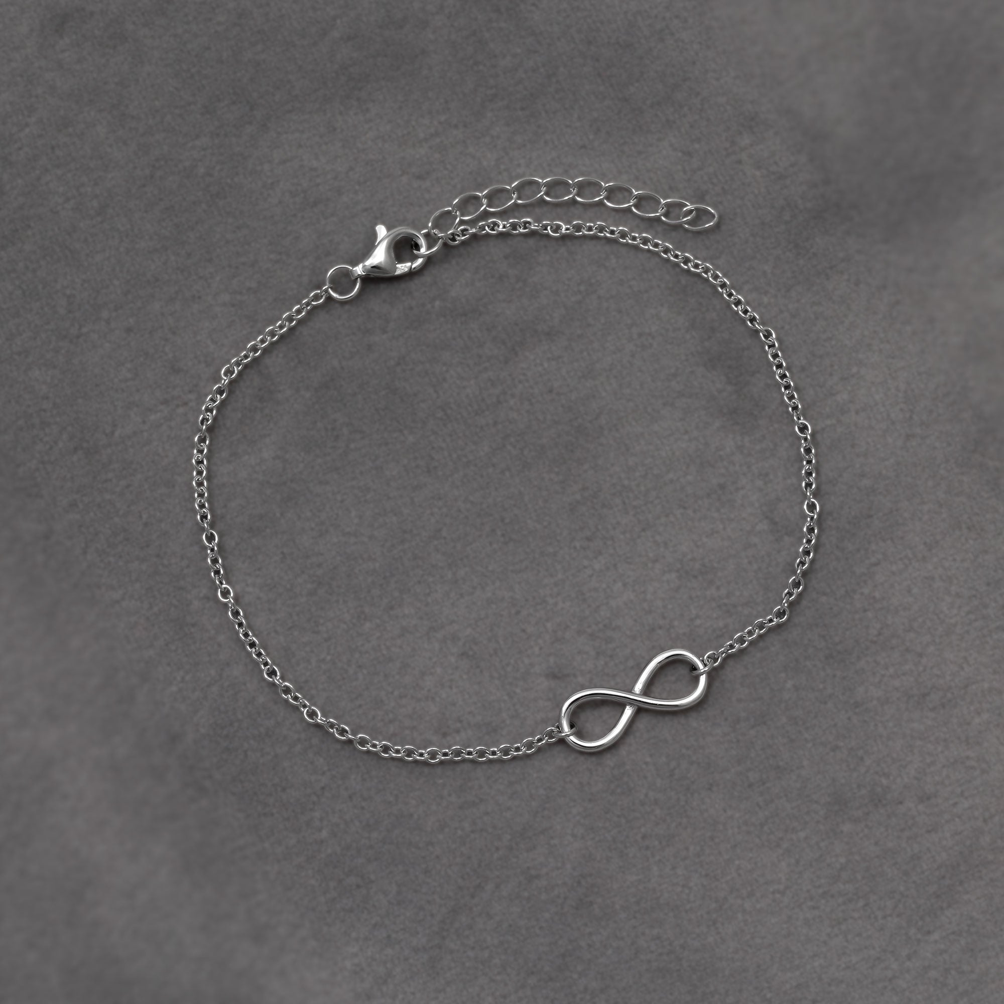 Delicate sterling silver bracelet featuring a polished infinity symbol centerpiece on a fine adjustable chain, embodying elegant and timeless design. placed on a grey velvet cloth.