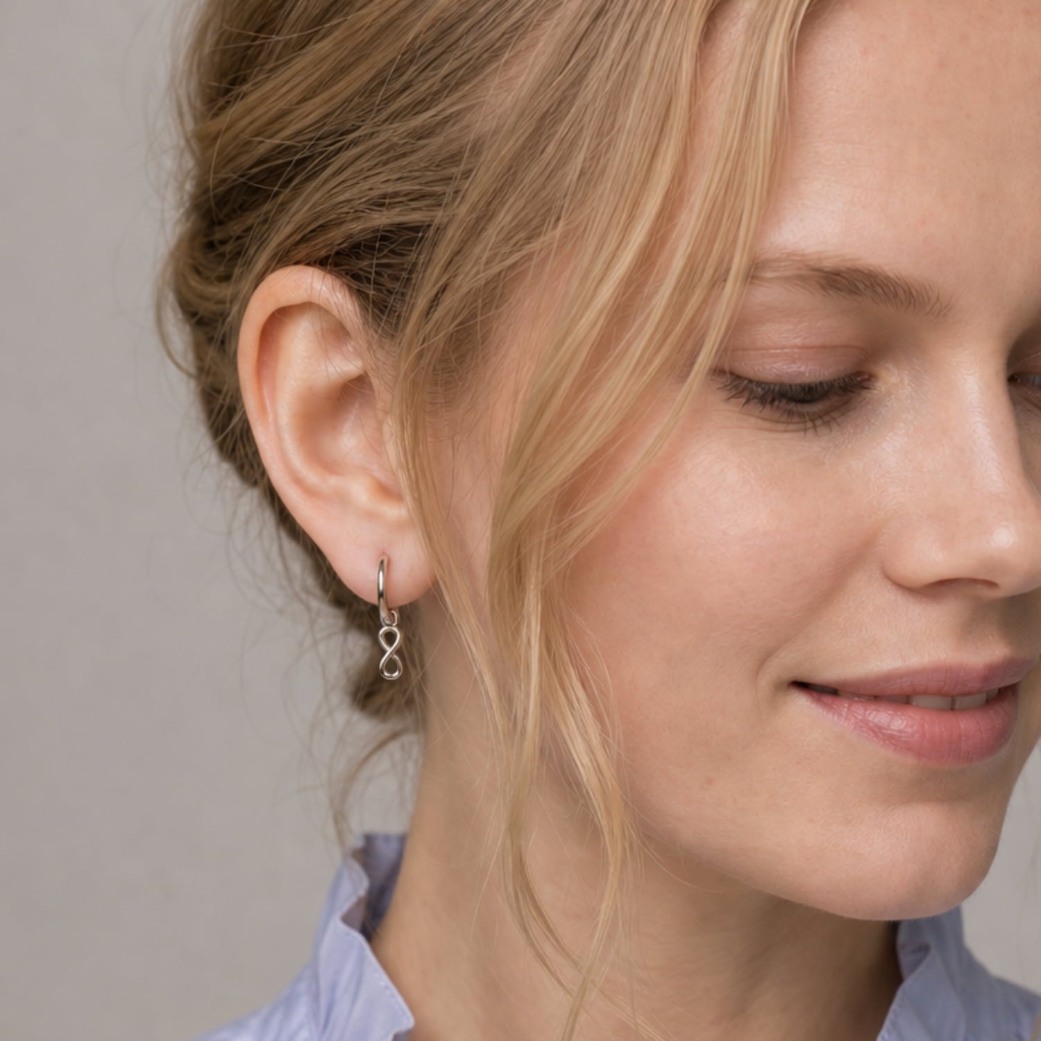 A close-up of a dangling sterling silver hoop earring featuring a minimalist infinity charm, worn on a woman's ear against a neutral background.