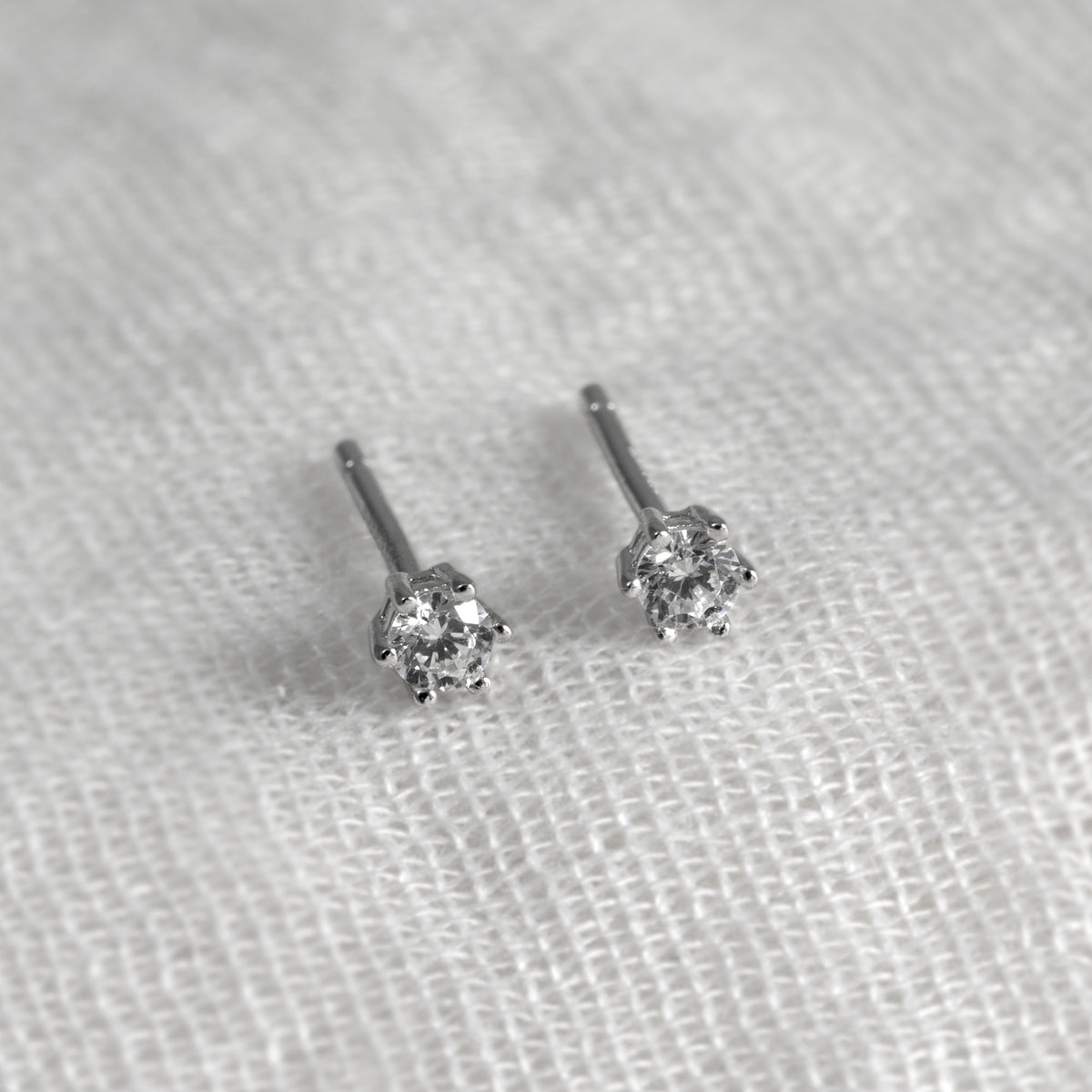 A pair of small silver stud earrings with clear cubic zirconia, resting on a textured white fabric background.