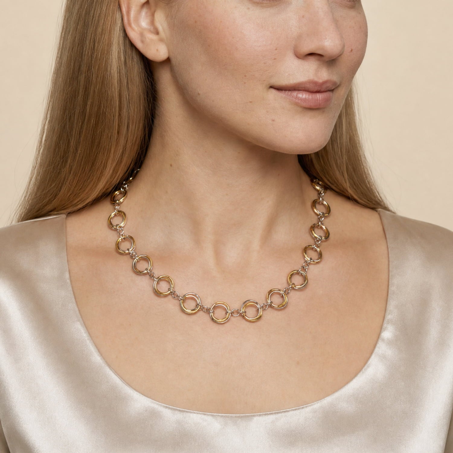 A close-up of a woman wearing a three tone ring-link necklace, paired with a champagne satin top against a warm beige background.