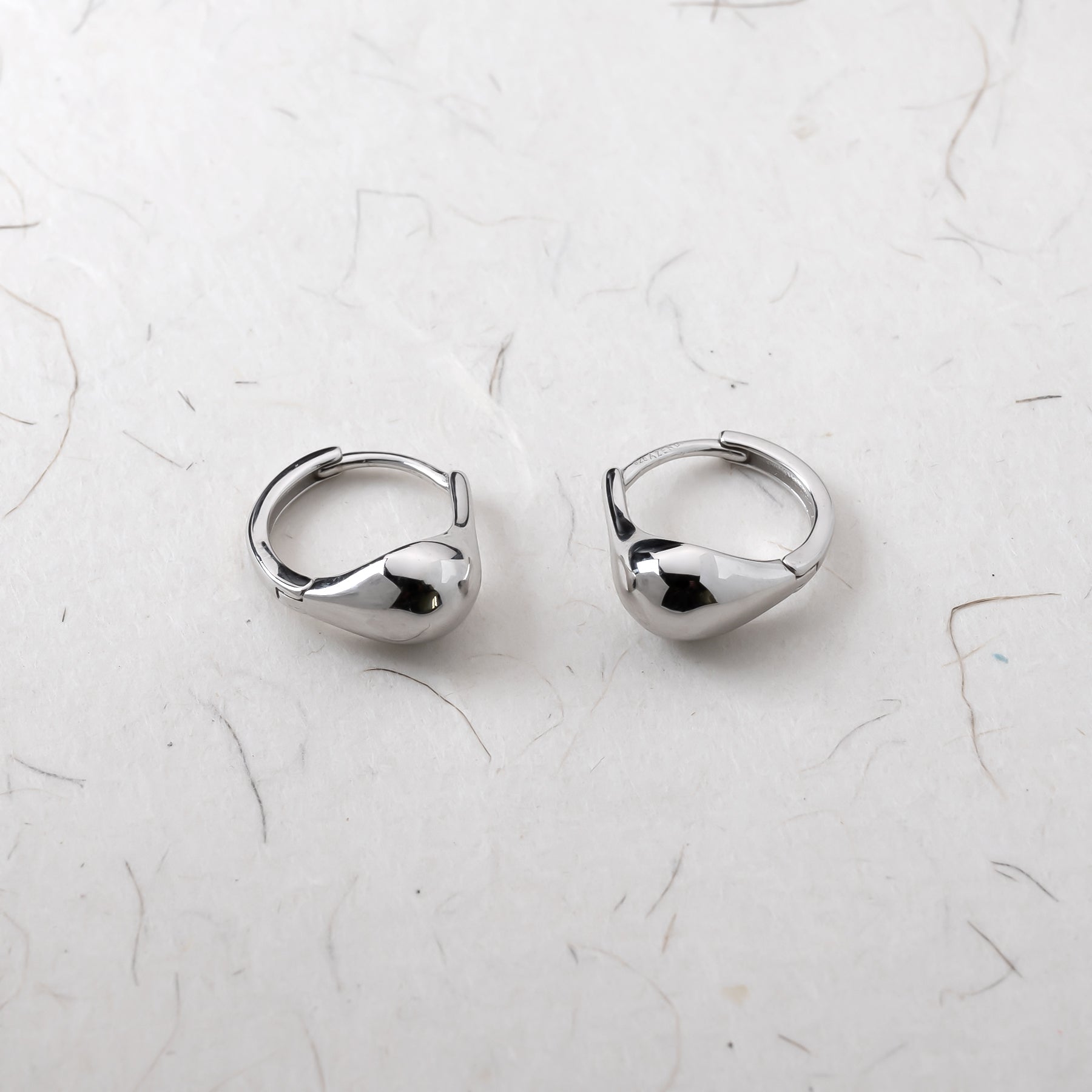 A pair of polished sterling silver teardrop hoop earrings with a smooth, sculptural design resting on a textured white surface.
