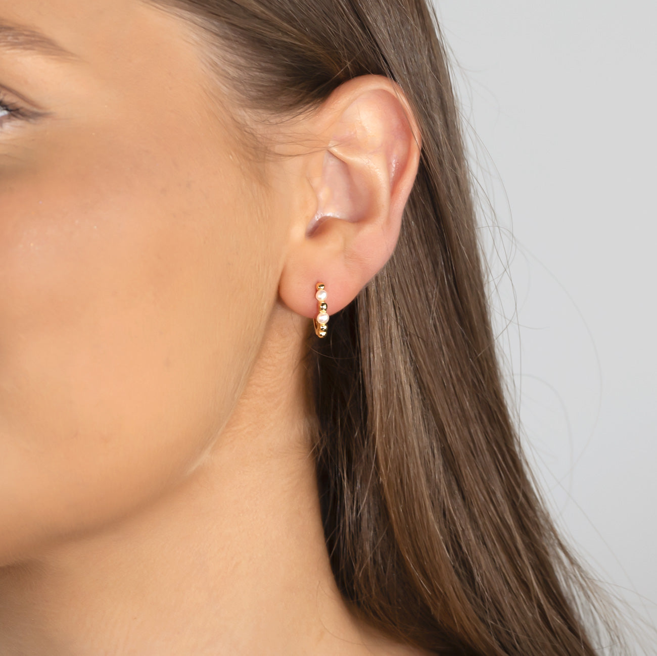 Close-up of a woman wearing a small sterling silver hoop earring featuring alternating pearls and beads, showcasing a delicate and sculptural design by Azendi.