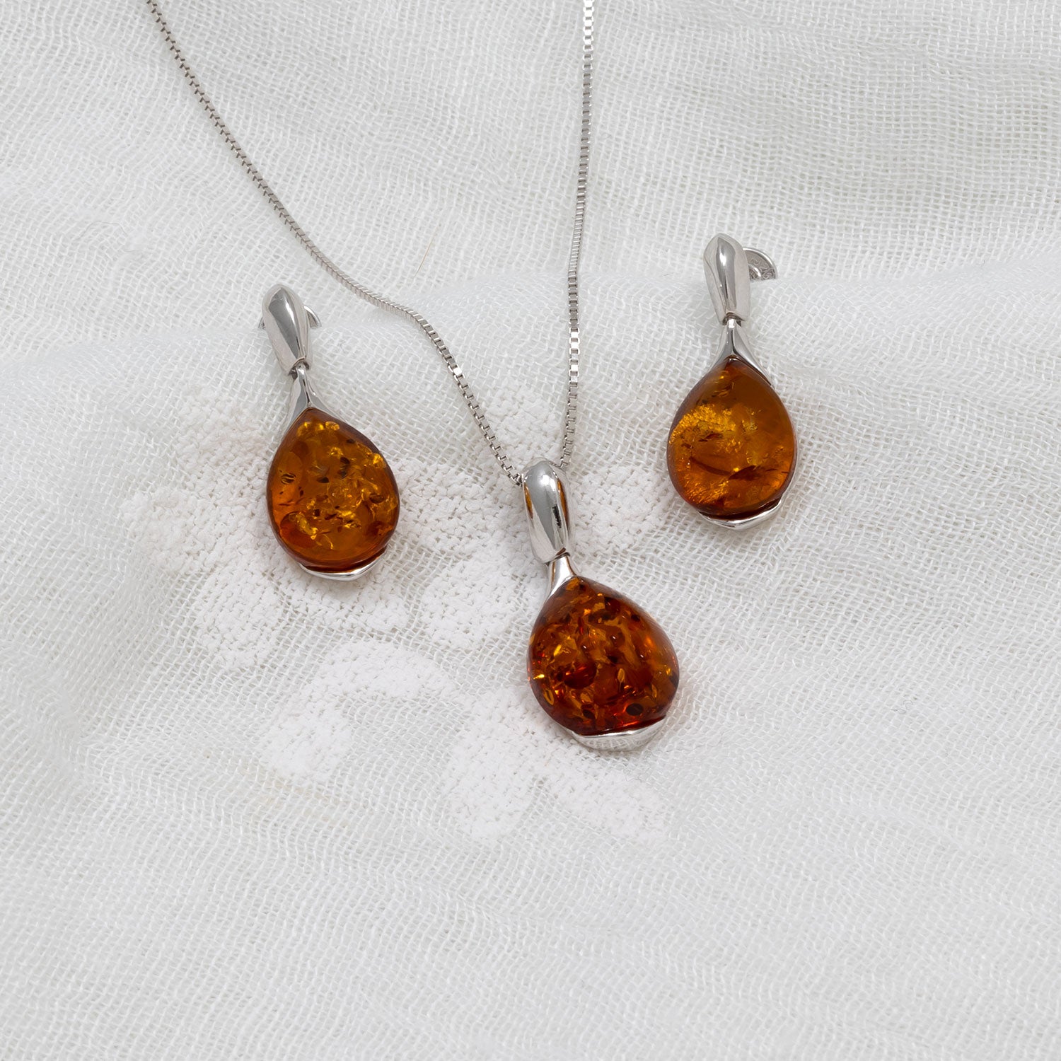 Pair of sterling silver earrings featuring amber droplets with natural inclusions, displayed alongside a matching amber pendant necklace on textured white fabric.