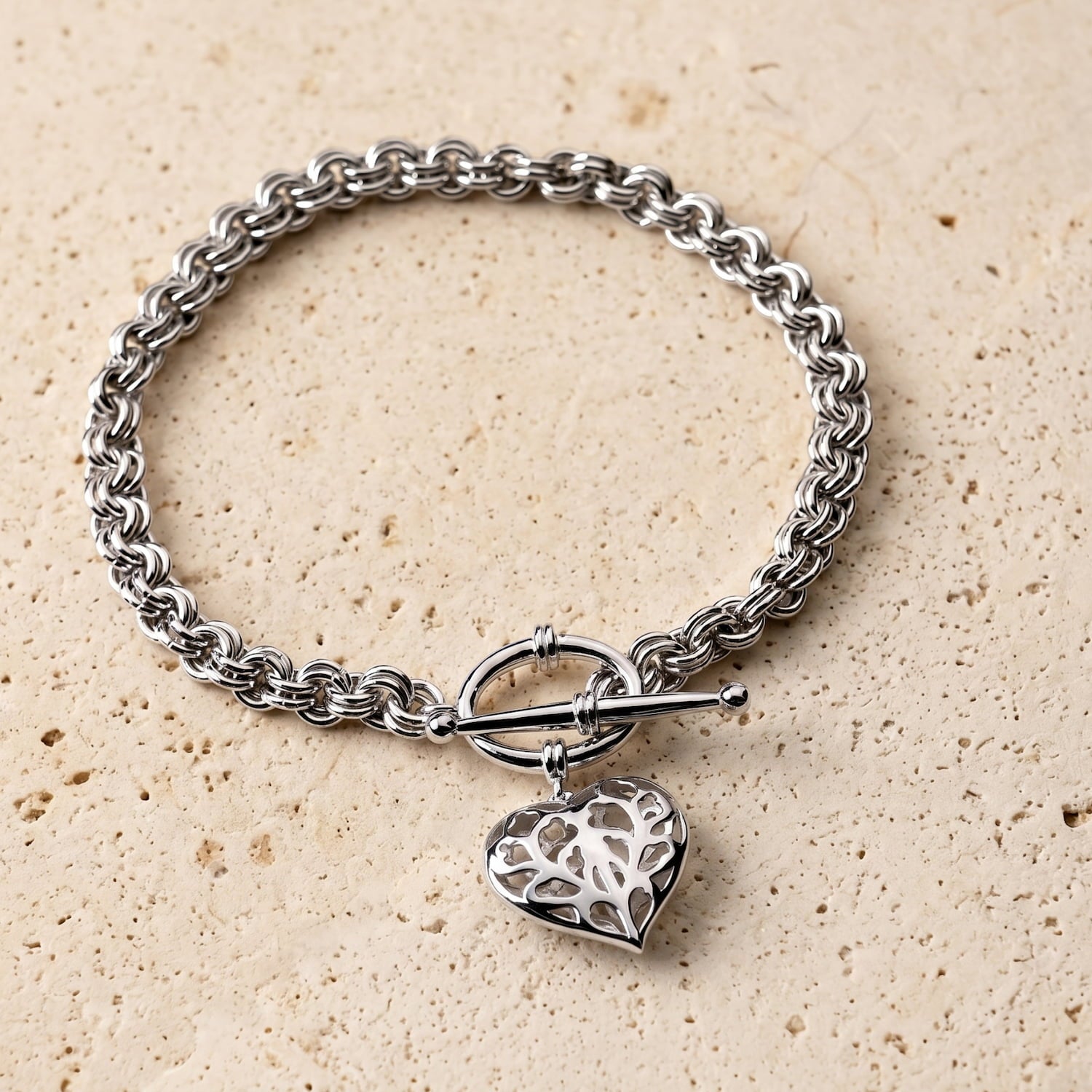 Sterling silver bracelet with a sculptural link chain and a hanging openwork heart charm, secured by a T-bar clasp, showcasing elegant British silver jewellery design.