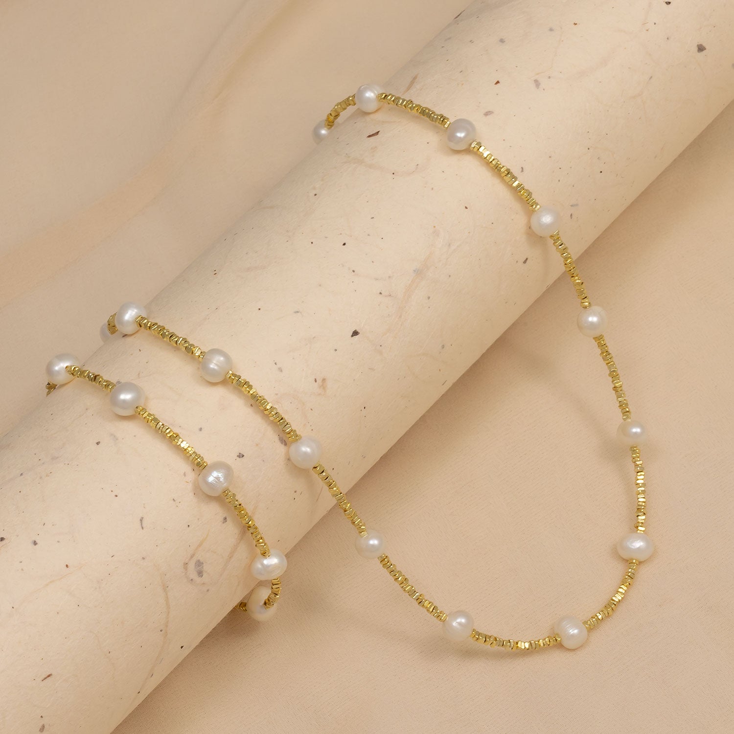 Elegant gold vermeil necklace featuring cubed beads interspersed with lustrous freshwater pearls, displayed on a soft beige textured surface.