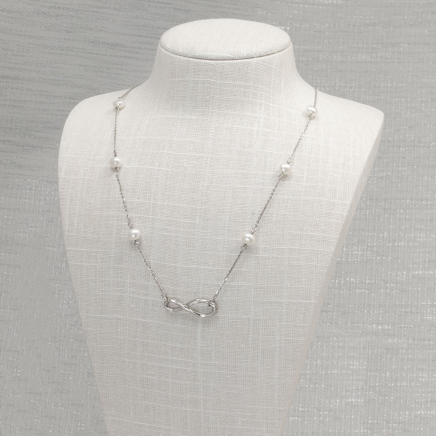 Sterling silver necklace featuring an infinity motif centerpiece with evenly spaced freshwater pearls along a delicate chain, displayed on a neutral fabric bust.