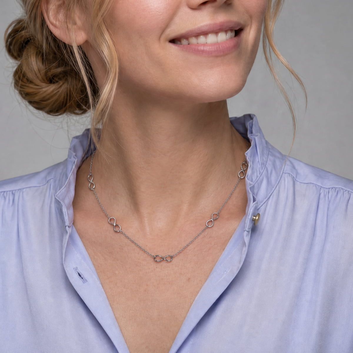 A close-up of a woman wearing a delicate silver chain necklace with small interlocking infinity motifs, styled with a light blue button-down shirt.