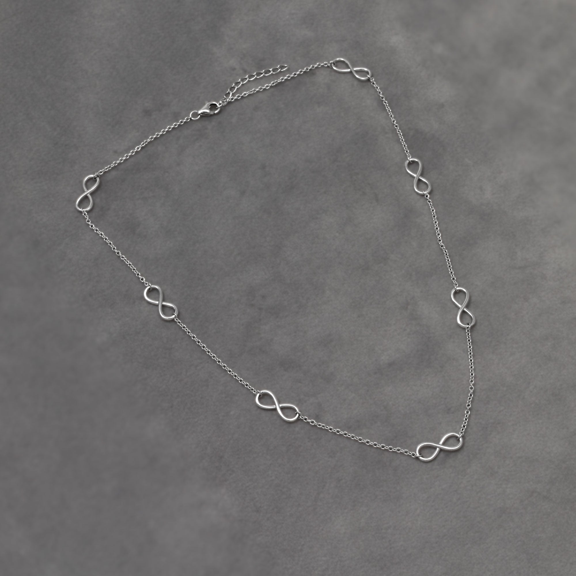 Delicate sterling silver necklace featuring multiple small infinity symbols spaced evenly along an adjustable chain, showcasing a minimalist and elegant design. Placed on a grey velvet cloth.