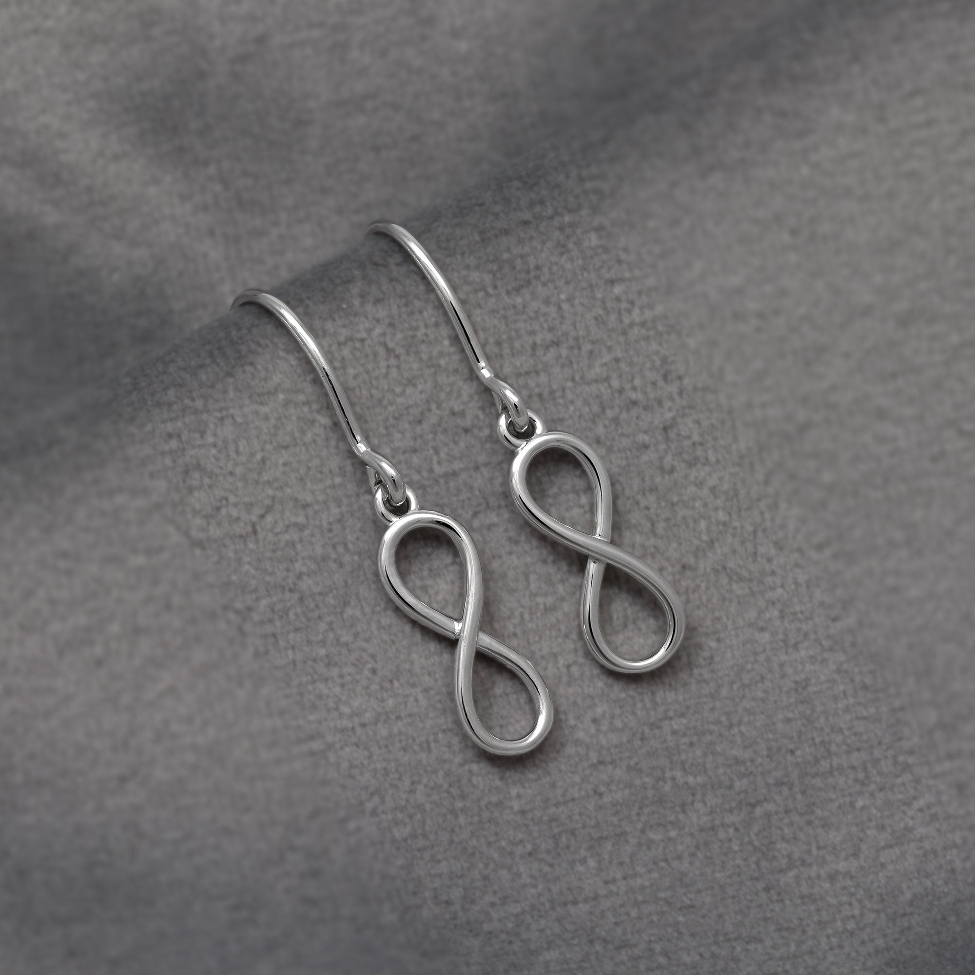 Pair of slender sterling silver drop earrings featuring a smooth infinity symbol suspended from simple curved hooks, embodying elegant and modern silver jewellery design. Placed on a grey velvet cloth.