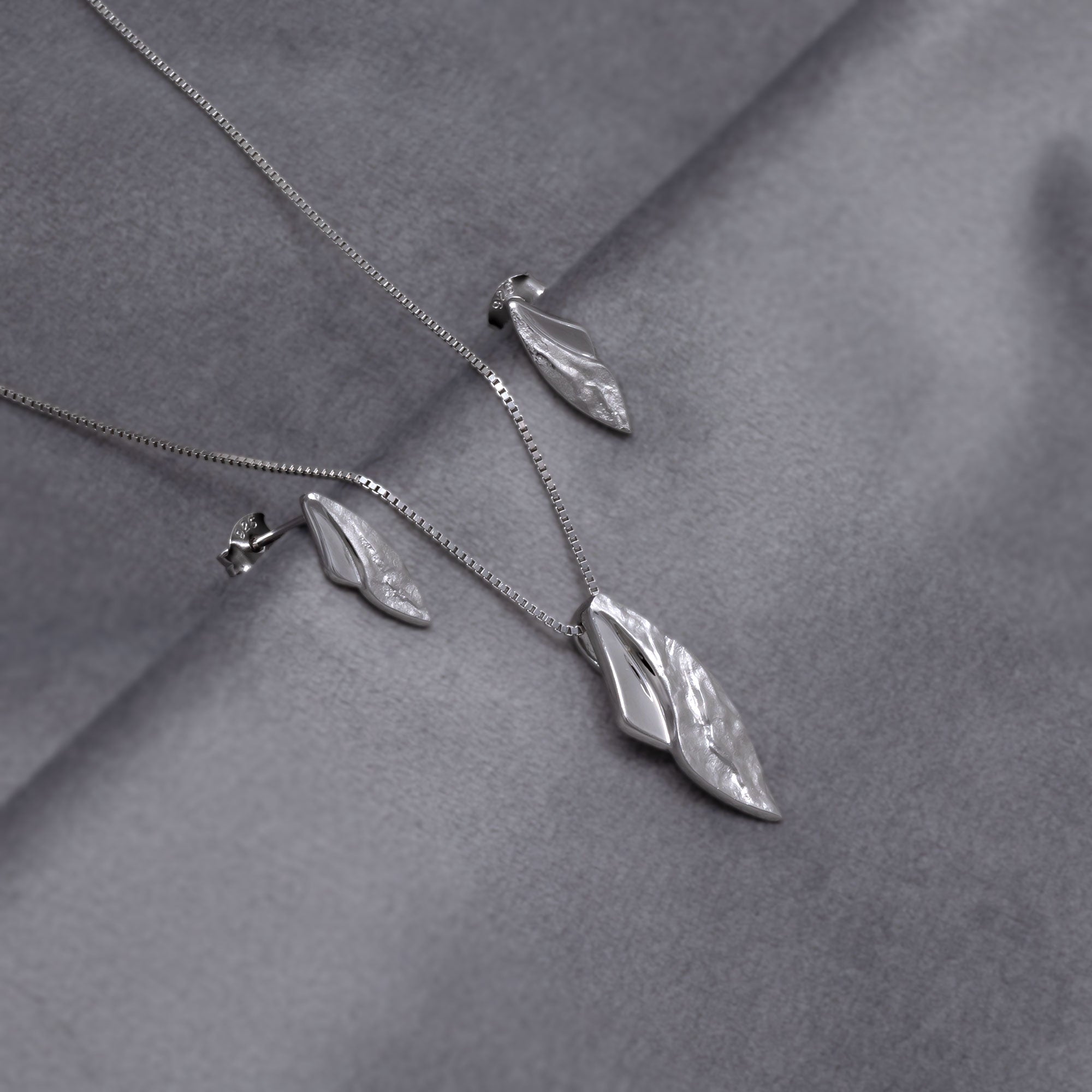 Sterling silver pendant and matching stud earrings combining curved polished and textured surfaces on a delicate chain, showcasing contemporary sculptural design in fine silver jewellery. Placed on a grey velvet cloth.