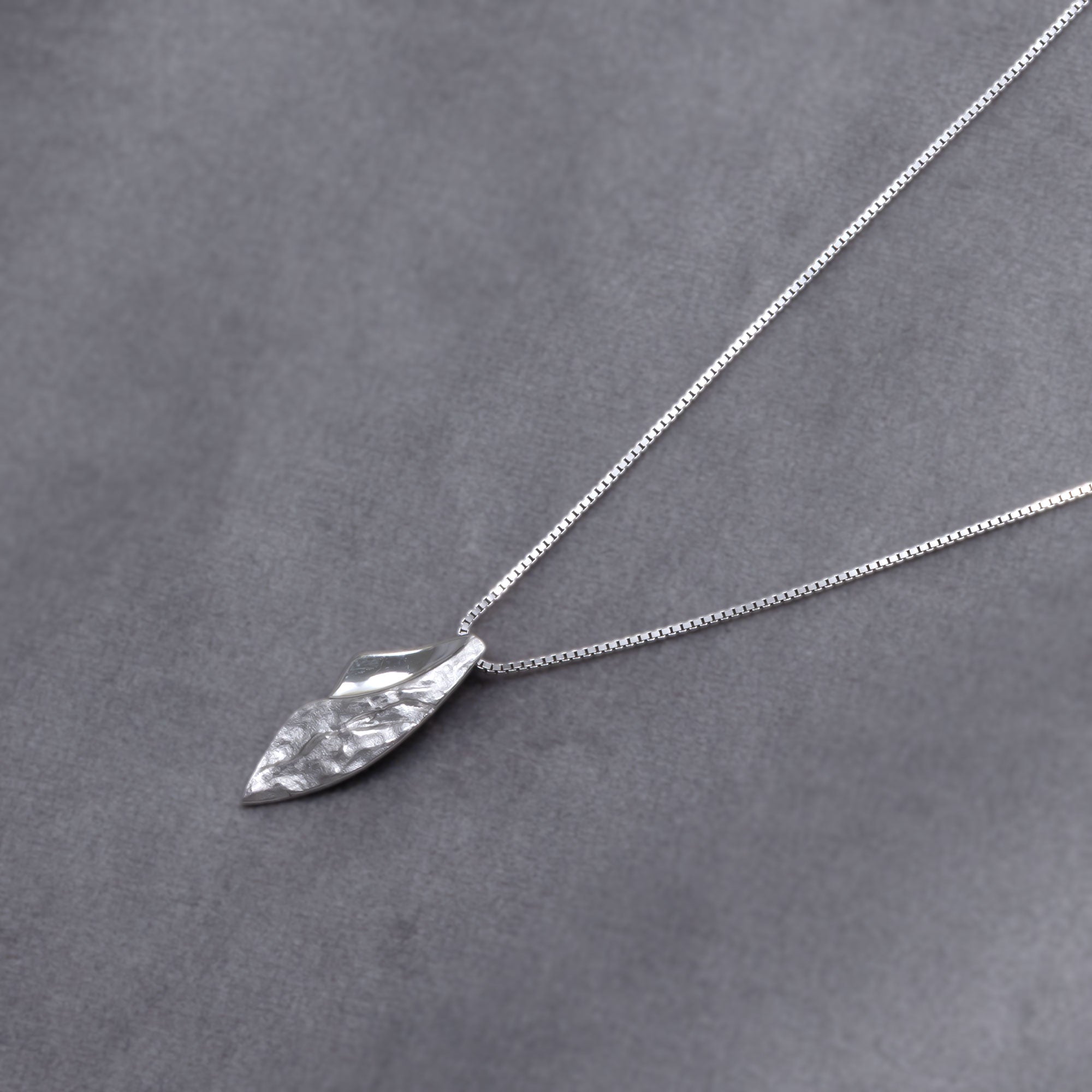 Sterling silver pendant combining curved polished and textured surfaces on a delicate chain, showcasing contemporary sculptural design in fine silver jewellery. Placed on a grey velvet cloth.