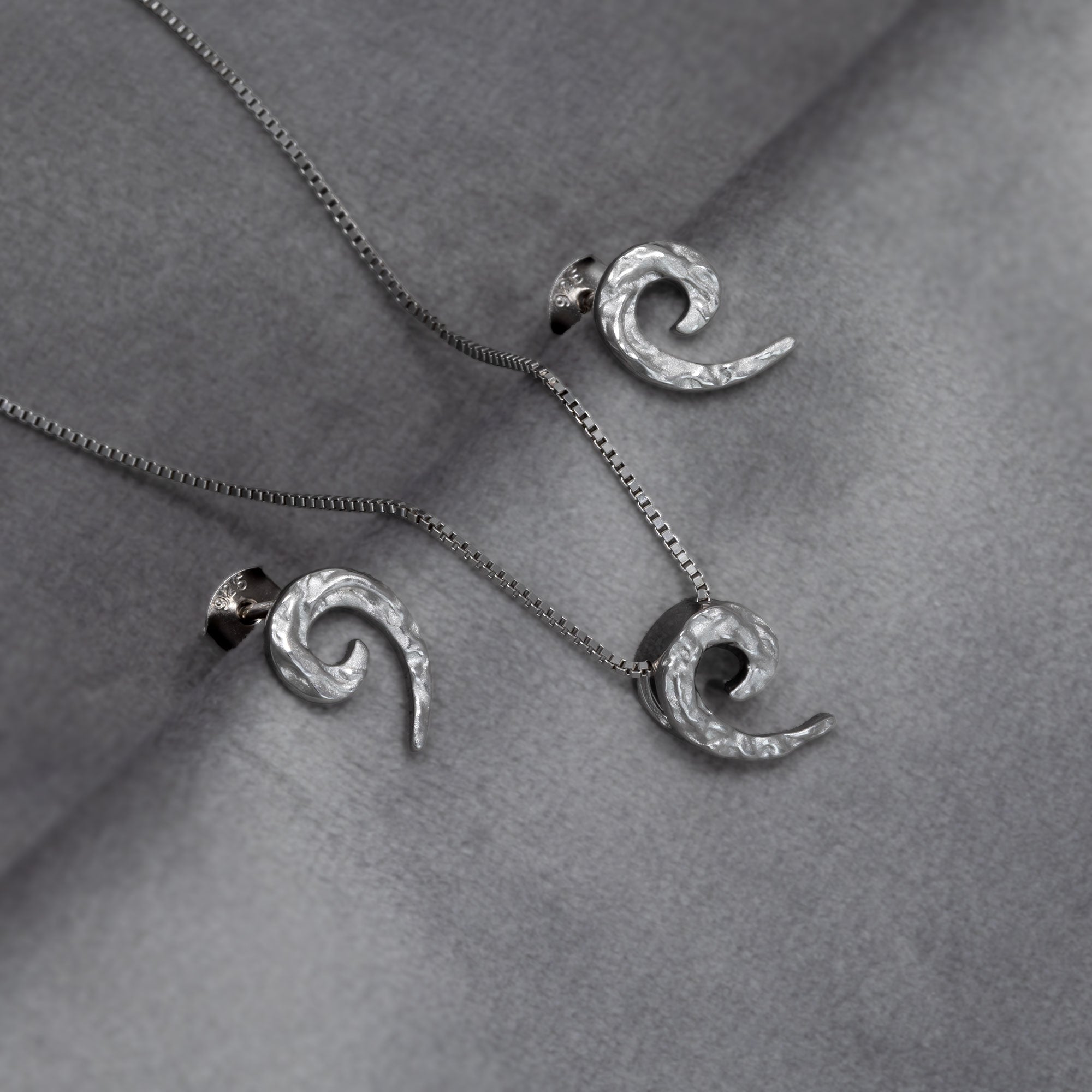 Sterling silver pendant and stud earrings with a textured curling horn design suspended on a delicate box chain, exemplifying sculptural elegance in contemporary silver jewellery. Placed on a grey velvet cloth.