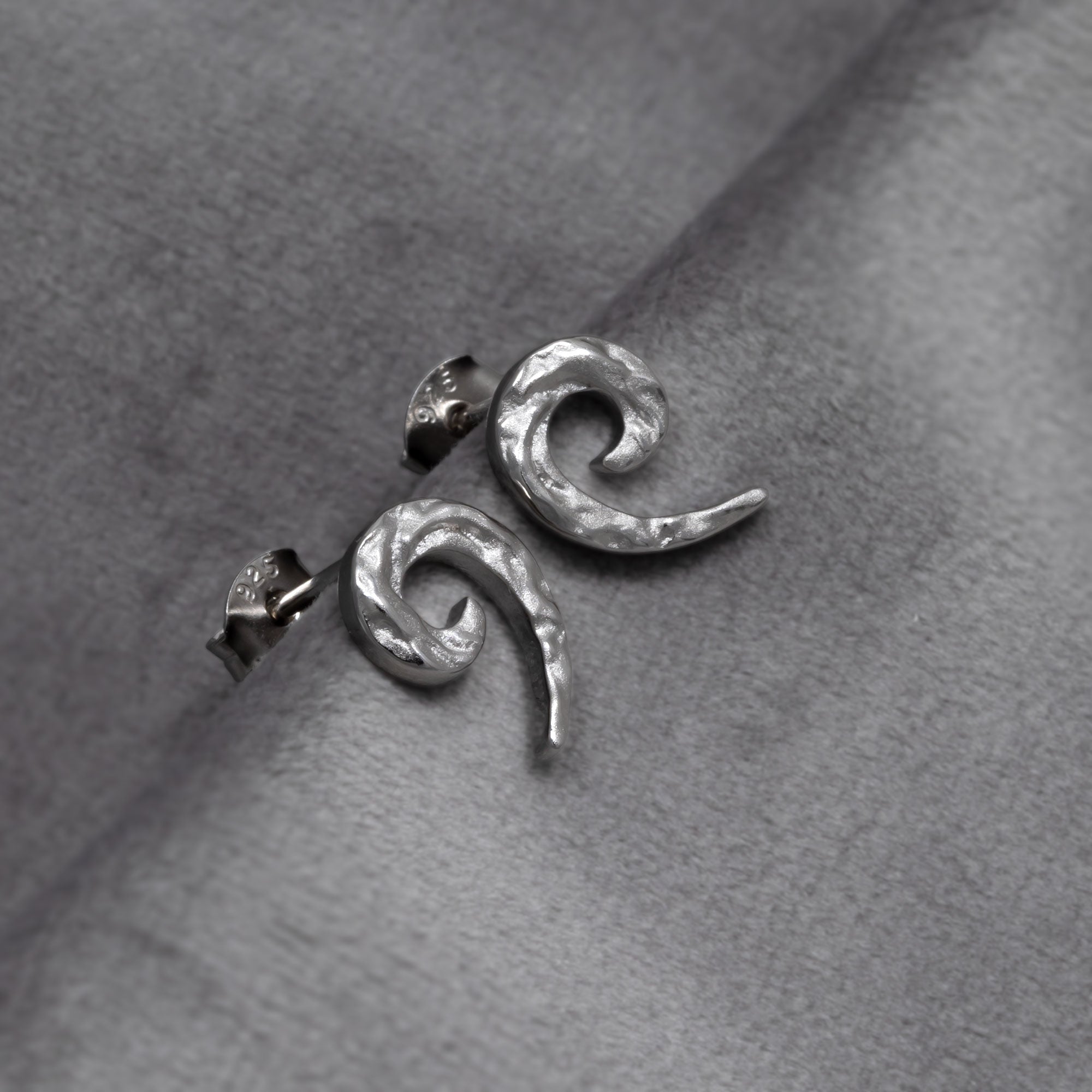 Pair of textured sterling silver stud earrings featuring a sculptural curling horn shape with a hammered finish, exemplifying refined contemporary silver jewellery. Placed on a grey velvet cloth.