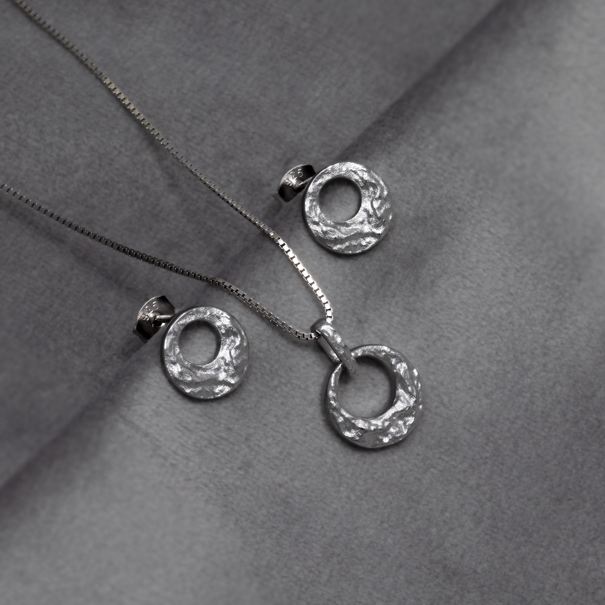 Sterling silver necklace featuring a textured circular pendant with a sculptural, hammered finish, showcasing contemporary British silver jewellery design. Placed on a grey velvet cloth. laced on a grey velvet cloth.