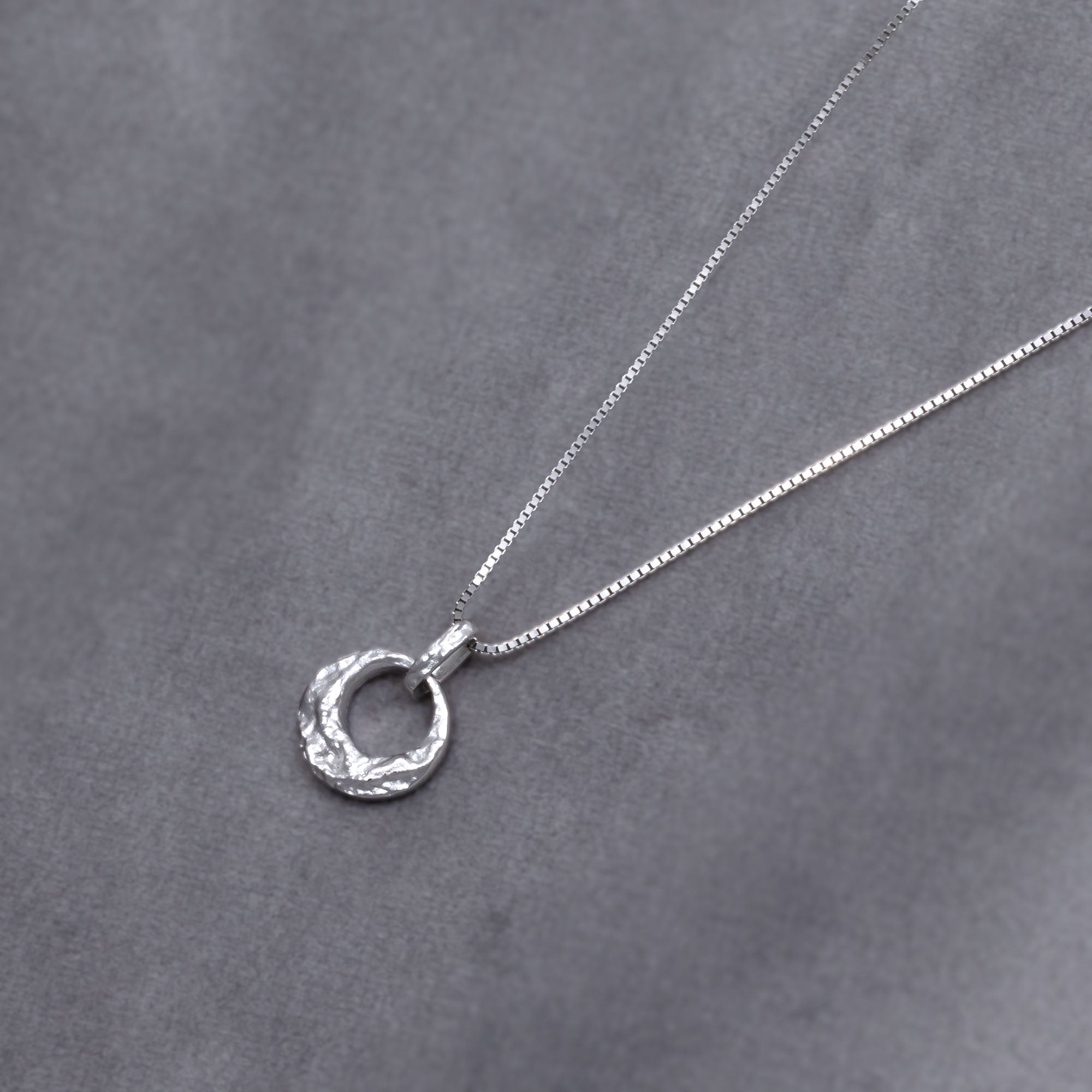 Sterling silver necklace featuring a textured circular pendant with a sculptural, hammered finish, showcasing contemporary British silver jewellery design. Placed on a grey velvet cloth.