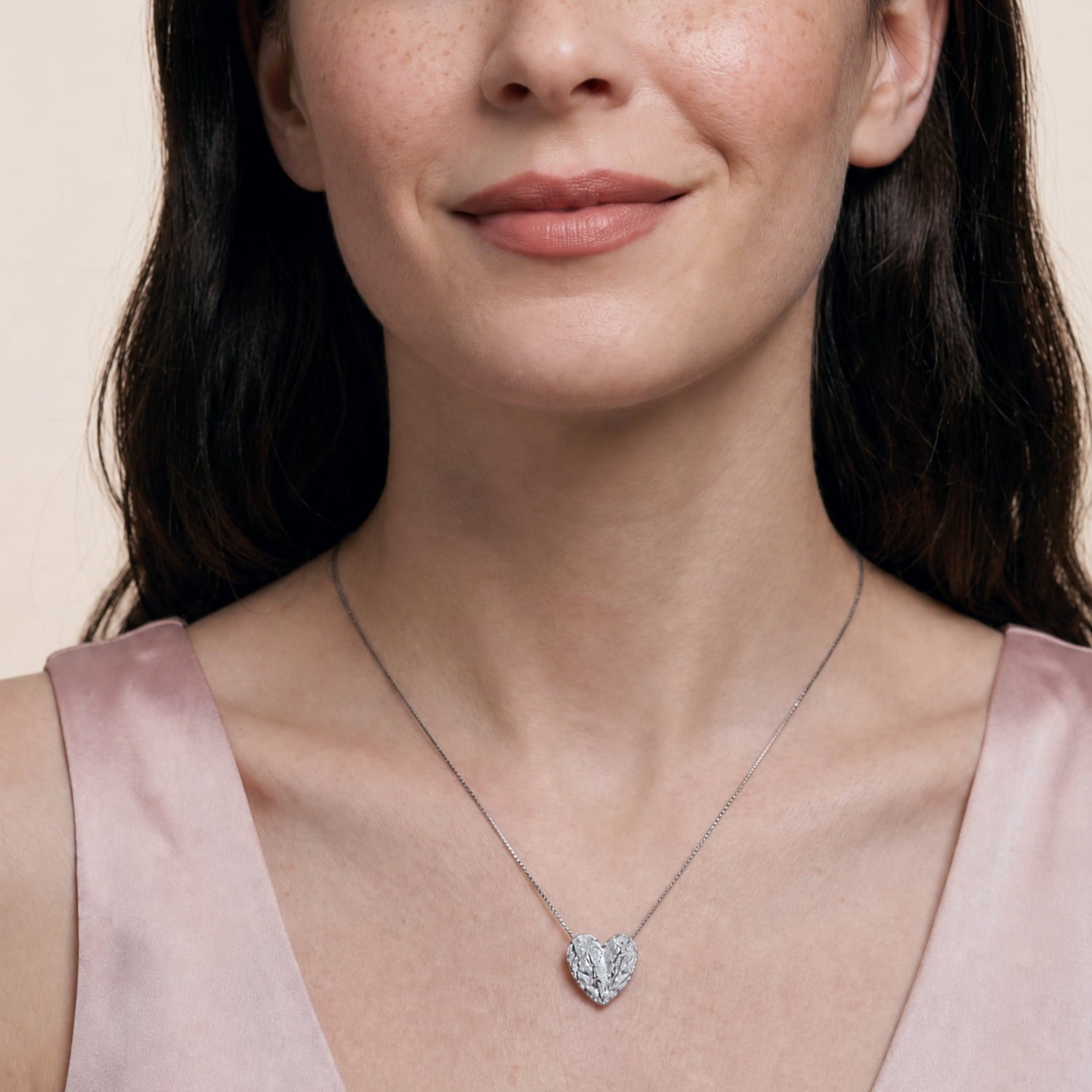 A close-up of a woman wearing a textured silver heart-shaped pendant necklace, styled with a light pink satin top against a soft beige background.