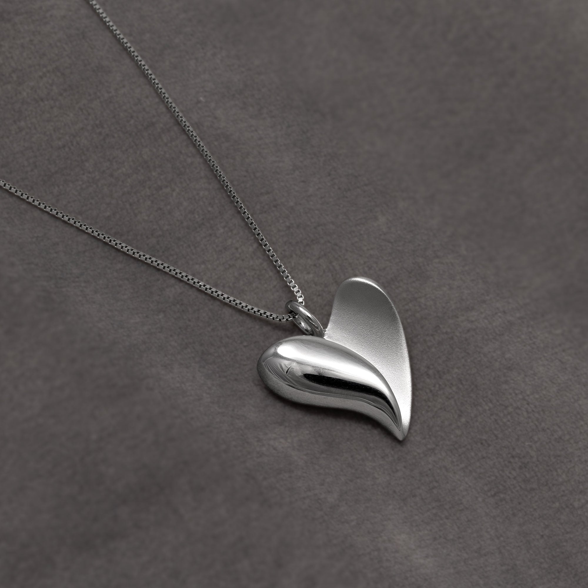 A polished silver pendant necklace featuring a heart shape on a delicate silver chain, displayed on a textured dark grey background.