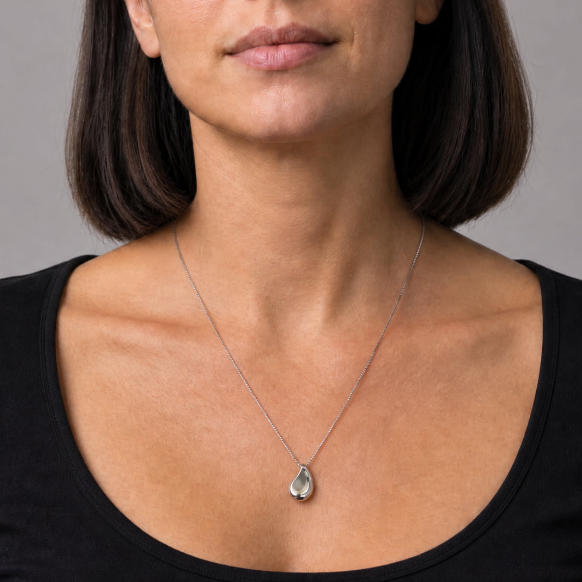 A close-up of a woman wearing a silver teardrop-shaped pendant, paired with a black scoop-neck top against a neutral gray background.