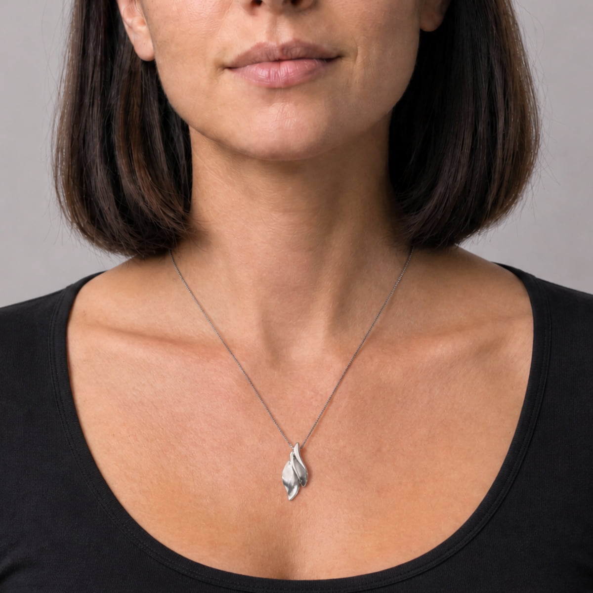 A close-up of a woman wearing a silver pendant necklace with a stylized leaf shape, paired with a black scoop-neck top against a neutral gray background.