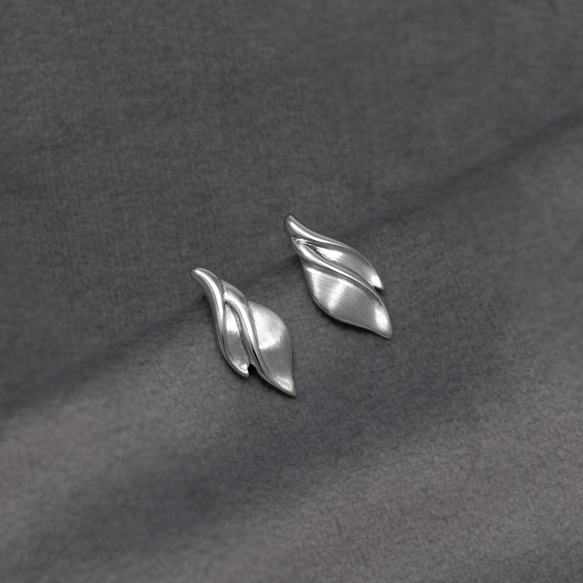 A pair of silver leaf-shaped stud earrings resting on a textured dark grey fabric background.