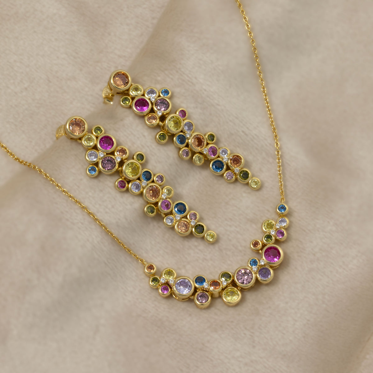 Gold vermeil drop earrings featuring a cluster of colourful bubble-like gemstones in varied hues, paired with a delicate matching necklace for a vibrant, sculptural look. Placed on a cream velvet cloth.