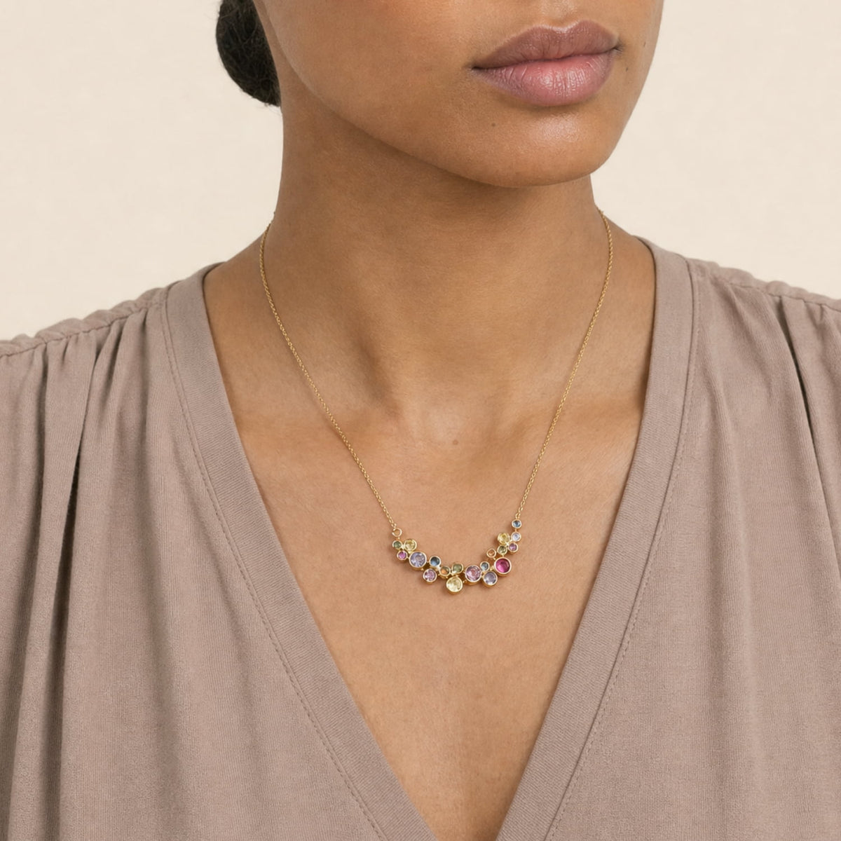 A close-up of a model wearing a gold vermeil necklace with a curved row of small, colorful stones at the center, paired with a taupe V-neck top against a light beige background.