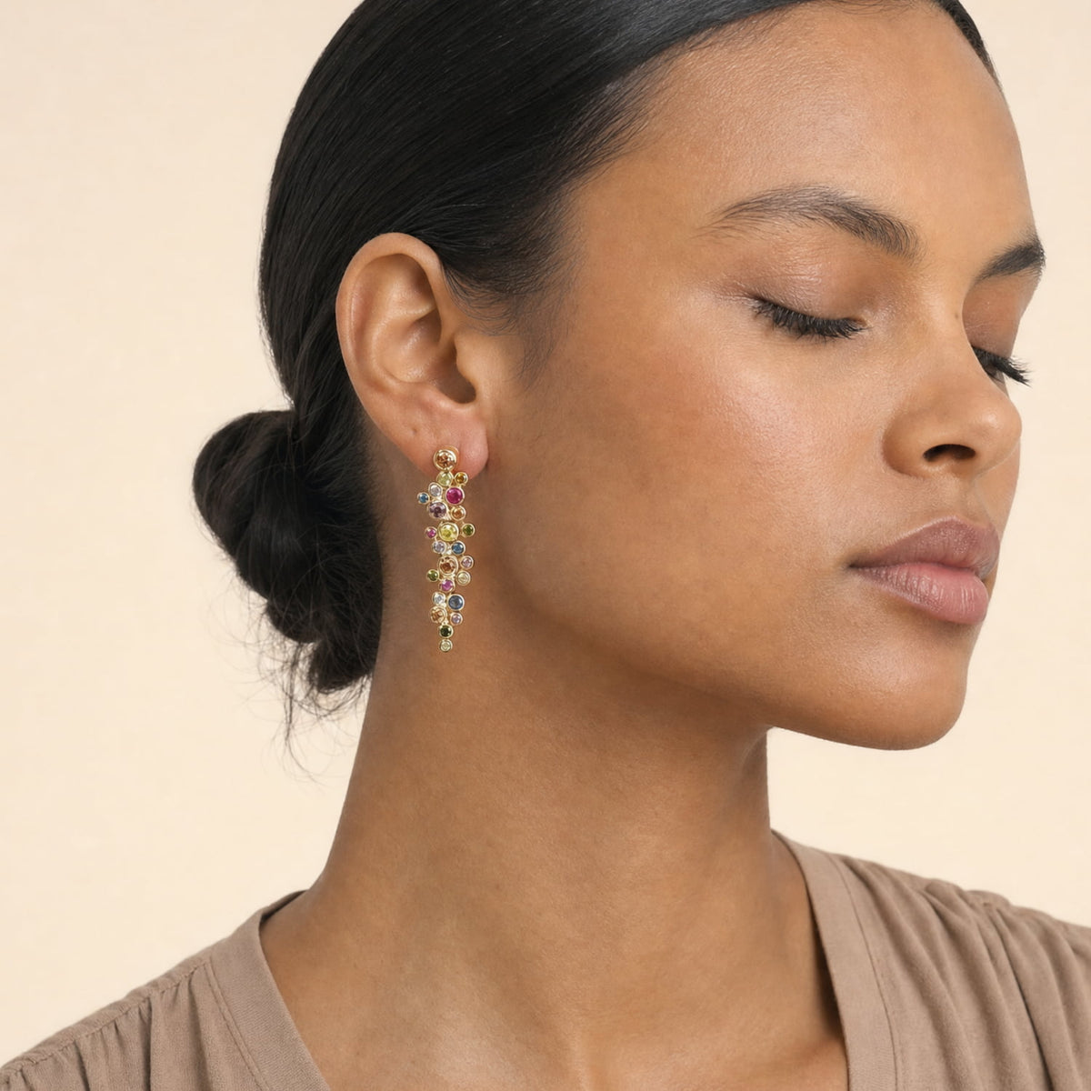 A close-up of a woman wearing a dangling multicolor cubic zirconia earring with yellow gold vermeil, set against a warm beige background.