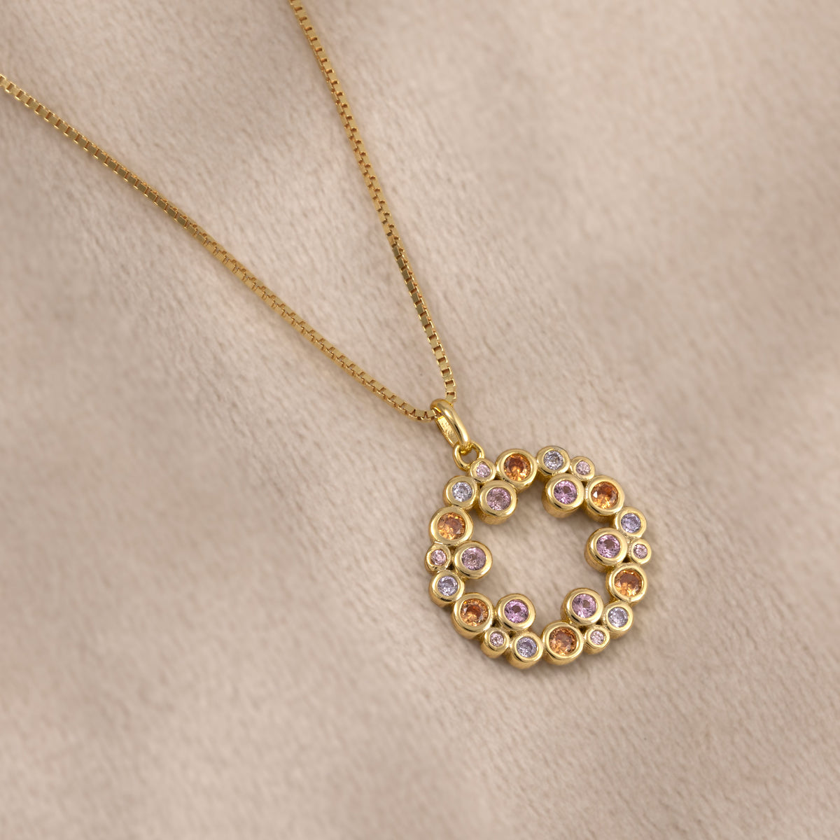 A gold vermeil circular pendant featuring colourful pink, purple, and orange bubble-shaped gemstones in a sculptural cluster design by Azendi. Placed on a cream velvet cloth.
